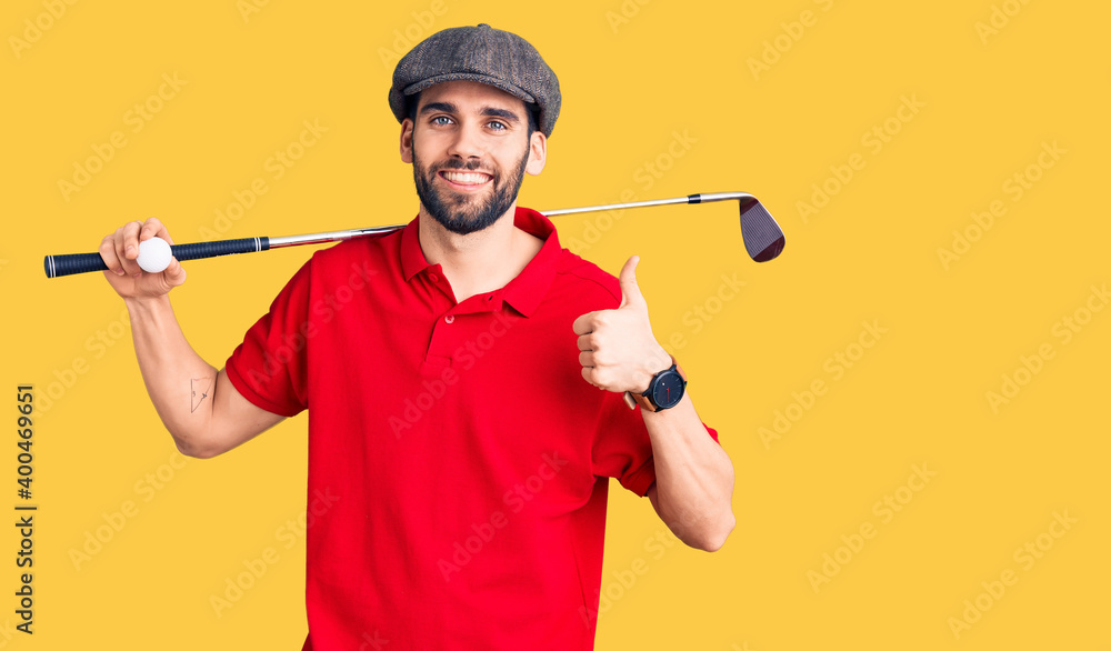 Young handsome man with beard playing golf holding club and ball smiling happy and positive, thumb up doing excellent and approval sign Young handsome man with beard playing golf holding club and ball smiling happy and positive, thumb up doing excellent and approval sign