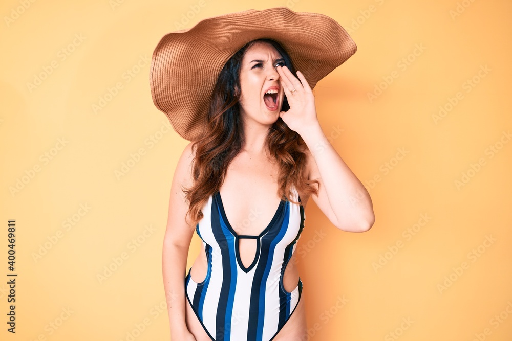 Young beautiful brunette woman wearing bikini and summer hat shouting and screaming loud to side with hand on mouth. communication concept. Young beautiful brunette woman wearing bikini and summer hat shouting and screaming loud to side with hand on mouth. communication concept.