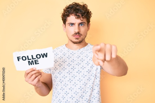 Young caucasian man with curly hair holding follow me message paper pointing with finger to the camera and to you, confident gesture looking serious © Krakenimages.com