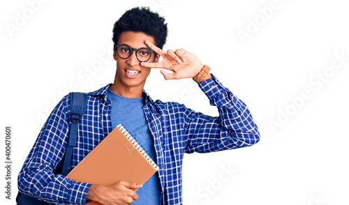 Young african american man wearing student backpack holding book doing peace symbol with fingers over face, smiling cheerful showing victory