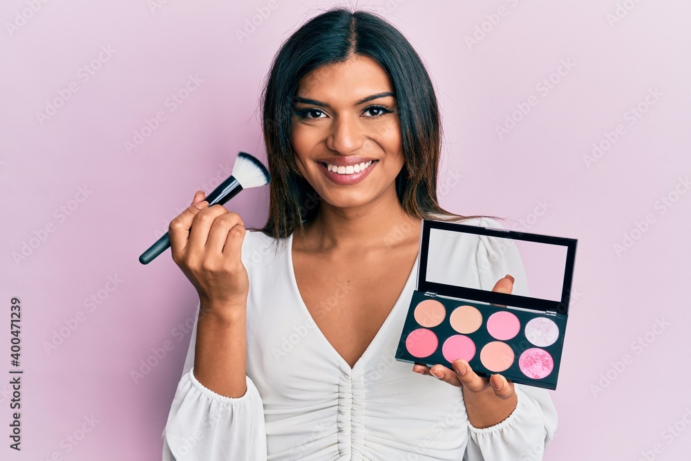 Young latin transsexual transgender woman holding makeup brush and blush palette smiling with a happy and cool smile on face. showing teeth. Young latin transsexual transgender woman holding makeup brush and blush palette smiling with a happy and cool smile on face. showing teeth.
