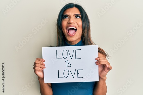 Young latin transsexual transgender woman holding paper with love is love message angry and mad screaming frustrated and furious, shouting with anger looking up. photo