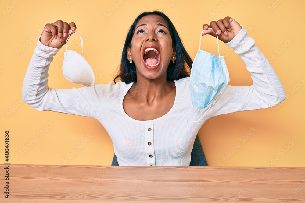 Young indian girl sitting on the table holding two diferent coronavirus protection mask angry and mad screaming frustrated and furious, shouting with anger looking up. Young indian girl sitting on the table holding two diferent coronavirus protection mask angry and mad screaming frustrated and furious, shouting with anger looking up.