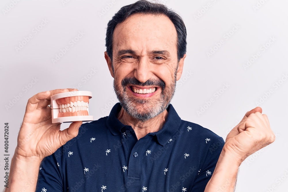 Middle age handsome man holding denture showing teeth over isolated white background screaming proud, celebrating victory and success very excited with raised arm Middle age handsome man holding denture showing teeth over isolated white background screaming proud, celebrating victory and success very excited with raised arm