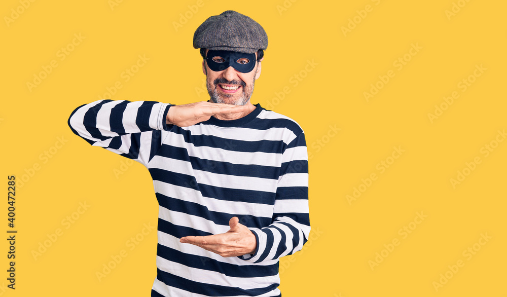 Middle age handsome man wearing burglar mask gesturing with hands showing big and large size sign, measure symbol. smiling looking at the camera. measuring concept. Middle age handsome man wearing burglar mask gesturing with hands showing big and large size sign, measure symbol. smiling looking at the camera. measuring concept.