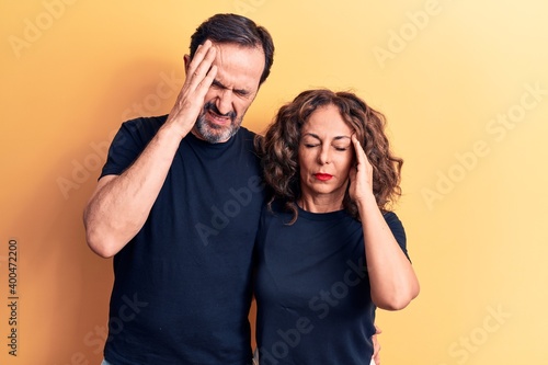 Middle age beautiful couple wearing t-shirt standing over isolated yellow background with hand on head, headache because stress. Suffering migraine.