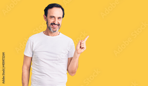 Middle age handsome man wearing casual t-shirt with a big smile on face, pointing with hand finger to the side looking at the camera.