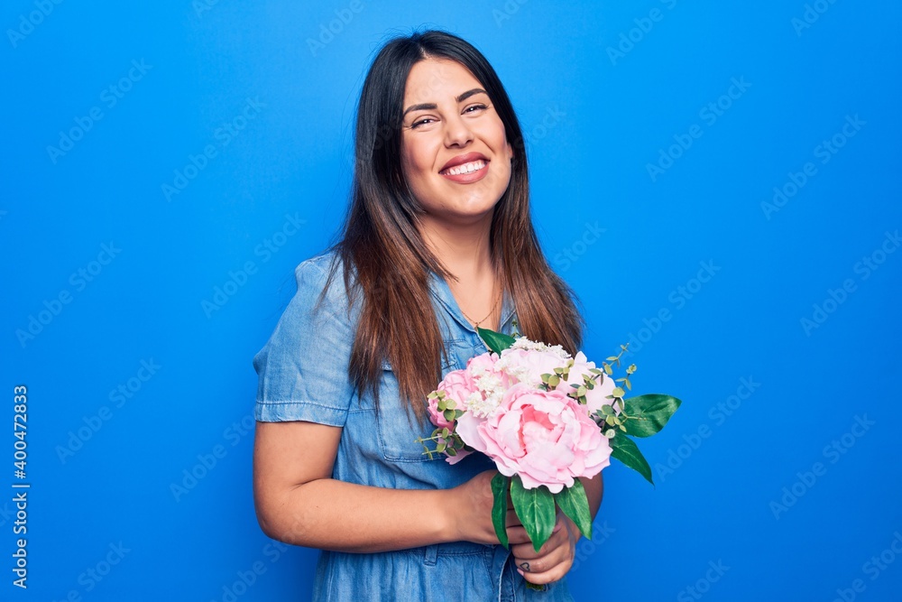 Young beautiful brunette woman holding bouquet of pink flowers over isolated blue background looking positive and happy standing and smiling with a confident smile showing teeth
