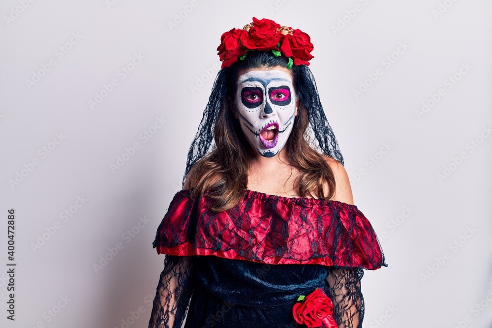 Young woman wearing day of the dead costume over white in shock face, looking skeptical and sarcastic, surprised with open mouth