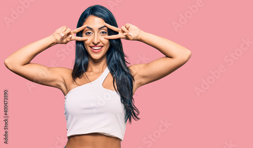 Beautiful hispanic woman wearing casual clothes doing peace symbol with fingers over face, smiling cheerful showing victory