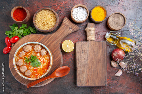 Above view of tomato meatballs soup with noodles in a brown bowl and different spices oil bottle onion garlic and cutting board on dark background