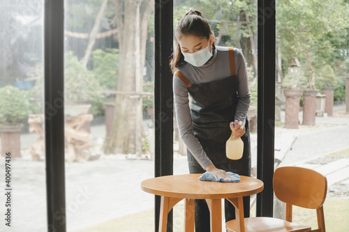 asian pretty waitress staff wearing protection face mask in apron cleaning table with disinfectant spray for protect infection coronavirus (covid-19) in cafe coffee shop restaurant. new normal concept