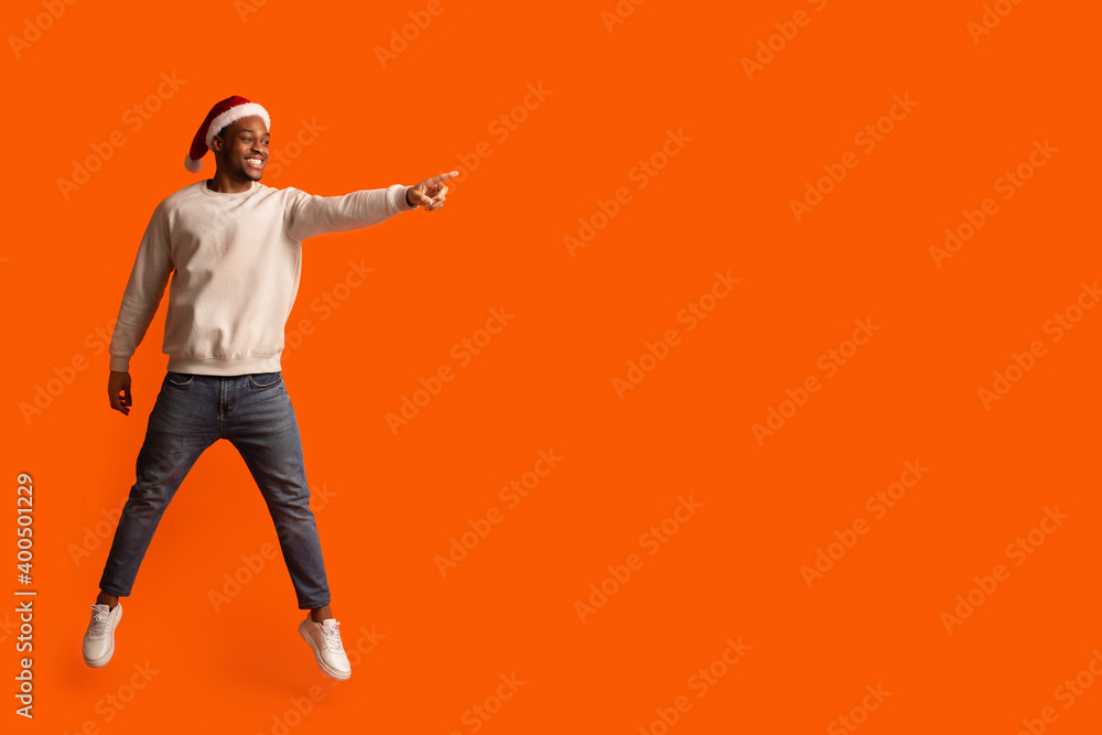 Christmas Promo. Excited Black Lady In Santa Hat Jumping And Pointing Aside Christmas Promo. Excited Black Lady In Santa Hat Jumping And Pointing Aside