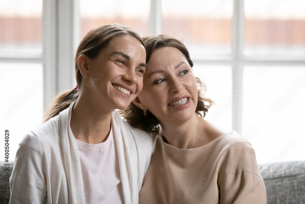 Close up head shot smiling dreamy grownup daughter and mother dreaming together, hugging, sitting on cozy couch at home, two generations visualizing future, enjoying leisure time, relaxing Close up head shot smiling dreamy grownup daughter and mother dreaming together, hugging, sitting on cozy couch at home, two generations visualizing future, enjoying leisure time, relaxing