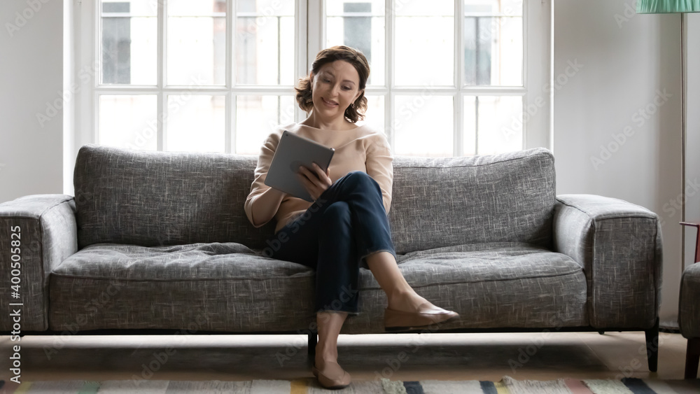 Smiling middle aged woman using tablet, surfing internet, sitting on cozy couch at home, happy mature female enjoying leisure time weekend with gadget, browsing apps, chatting or shopping online