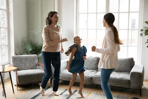 Excited little girl with mature grandmother and young mother dancing at home together, moving to favorite music, happy three generations of women having fun in modern apartment, enjoying leisure time