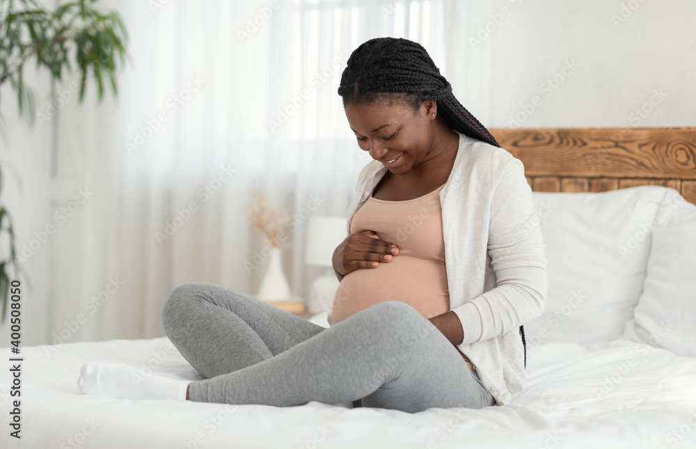 Happy Black Expectant Lady Enjoying Her Pregnancy, Sitting On Bed Touching Belly Happy Black Expectant Lady Enjoying Her Pregnancy, Sitting On Bed Touching Belly