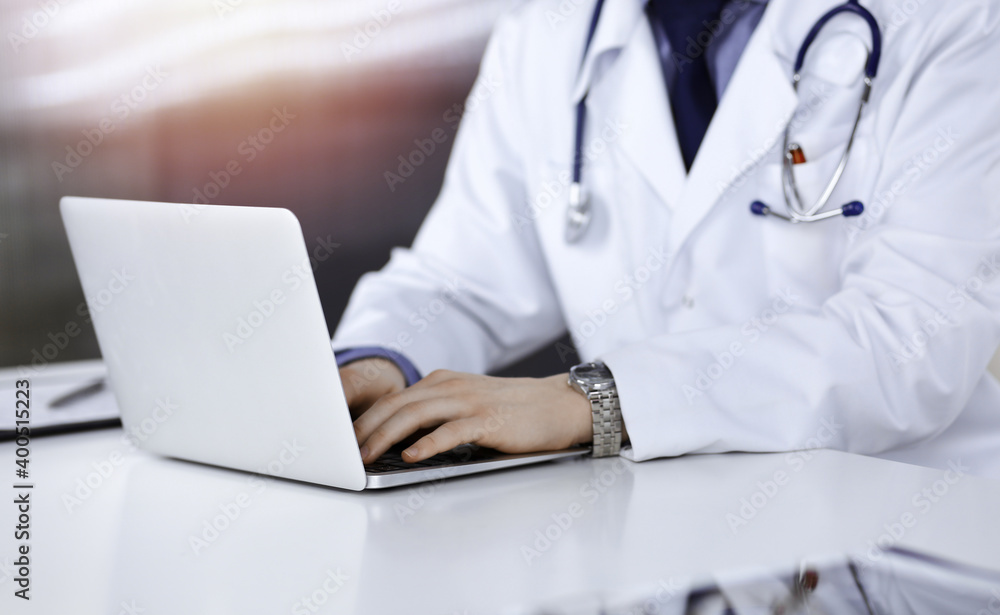 Unknown male doctor sitting and working with laptop in a darkened clinic, glare of light on the background, close-upof hands Unknown male doctor sitting and working with laptop in a darkened clinic, glare of light on the background, close-upof hands