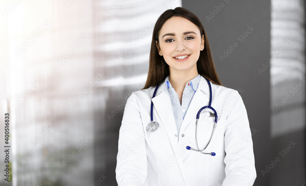 Smiling young woman-doctor is standing in sunny clinic indoors Smiling young woman-doctor is standing in sunny clinic indoors