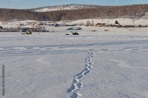 Human footprints lead to the rural shore along the frozen snow-covered surface of a large lake.