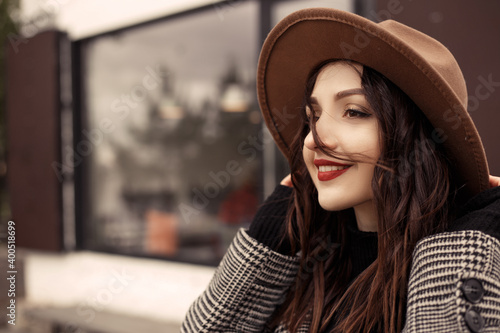 Close up shot charming woman standing outside near street cafe restaurant wear brown hat gray coat poloneck black sweater, looking away. Girl spend time autumn or spring, cold weather fresh air.  © Volodymyr