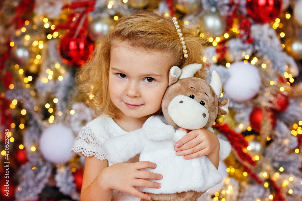 A little blonde girl in a white dress with a plush bull toy on the background of a Christmas tree and lights of garlands.