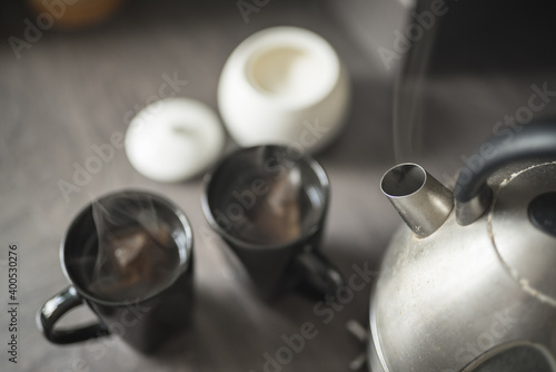 Close up of two black mugs with steaming water and teabags inside beside a boiling kettle, a spoon and a sugar bowl on a kitchen top in a House in Edinburgh, Scotland, United Kingdom