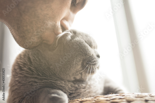 A man with a stubble kissing his British Short Hair cat in the head as she lies on a wicker stool near a patio door in a house in Edinburgh, Scotland, United Kingdom photo