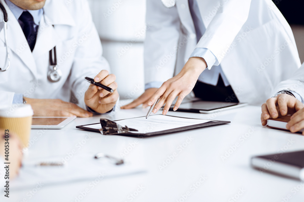 Group of unknown doctors are sitting at the desk and discussing medical treatment, using a clipboard, close-up. Team of physicians at work in a clinic. Medicine and healthcare concept Group of unknown doctors are sitting at the desk and discussing medical treatment, using a clipboard, close-up. Team of physicians at work in a clinic. Medicine and healthcare concept