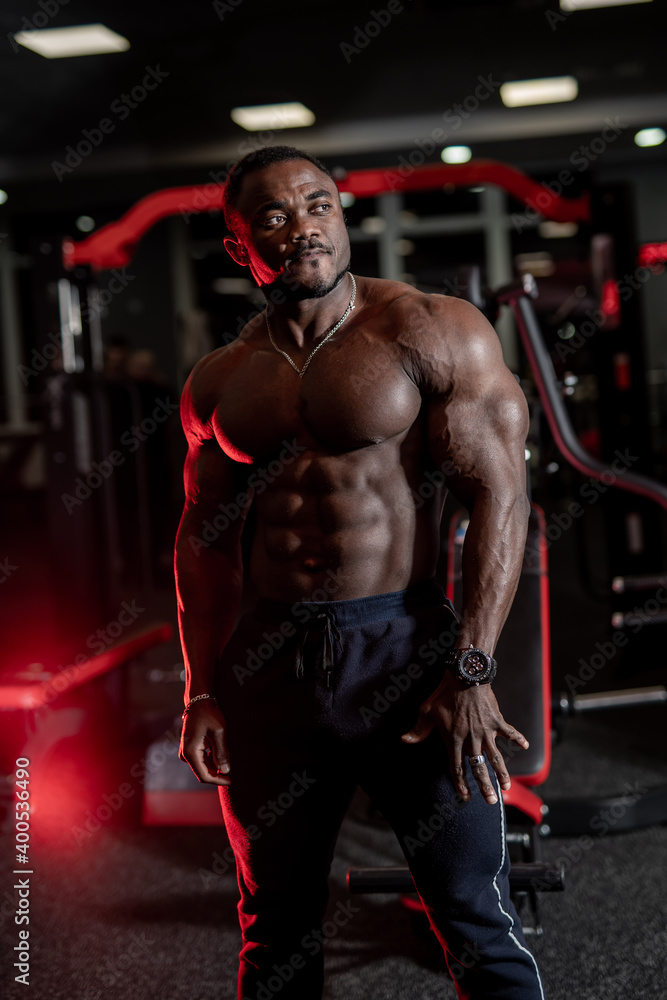 Portrait of an african american male fitness trainer. Bodybuilder shows biceps and press and looks at side gym background.