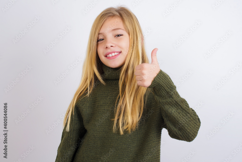 Caucasian kid girl wearing green knitted sweater against white wall doing happy thumbs up gesture with hand. Approving expression looking at the camera showing success. Caucasian kid girl wearing green knitted sweater against white wall doing happy thumbs up gesture with hand. Approving expression looking at the camera showing success.