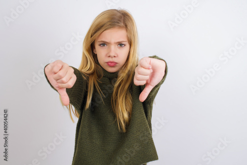 Wallpaper Mural Caucasian kid girl wearing green knitted sweater against white wall being upset showing thumb down with two hands. Dislike concept. Torontodigital.ca