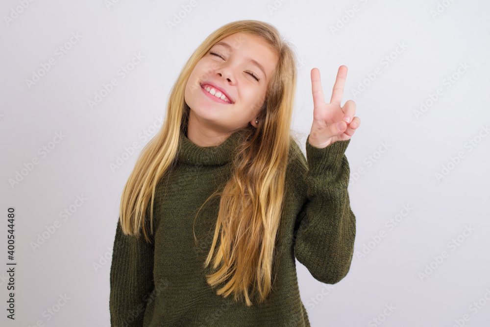 Caucasian kid girl wearing green knitted sweater against white wall smiling with happy face winking at the camera doing victory sign. Number two. Caucasian kid girl wearing green knitted sweater against white wall smiling with happy face winking at the camera doing victory sign. Number two.