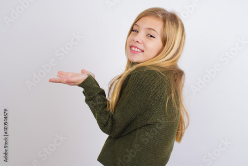Caucasian kid girl wearing green knitted sweater against white wall pointing aside with hands open palms showing copy space, presenting advertisement smiling excited happy