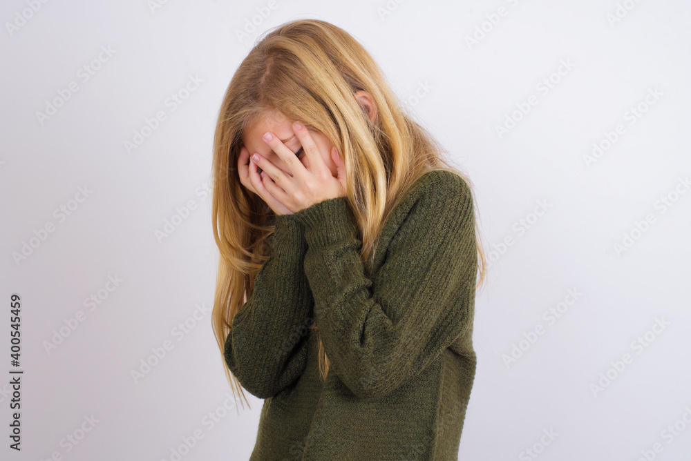 Sad Caucasian kid girl wearing green knitted sweater against white wall crying covering her face with her hands. Sad Caucasian kid girl wearing green knitted sweater against white wall crying covering her face with her hands.