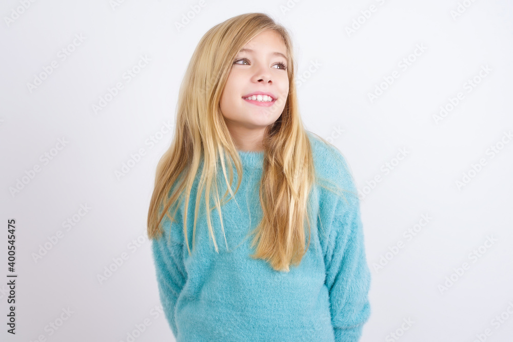 Cute Caucasian kid girl wearing blue knitted sweater against white wall with thoughtful expression, looks away keeps hands down bitting his lip thinks about something pleasant. Cute Caucasian kid girl wearing blue knitted sweater against white wall with thoughtful expression, looks away keeps hands down bitting his lip thinks about something pleasant.