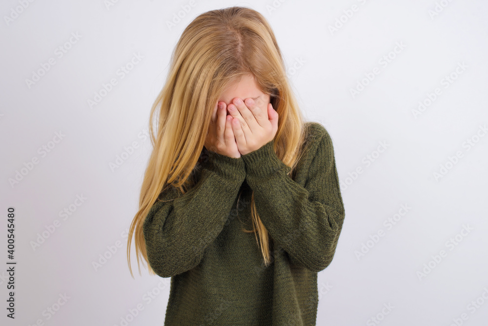 Caucasian kid girl wearing green knitted sweater against white wall covering her face with her hands, being devastated and crying. Sad concept Caucasian kid girl wearing green knitted sweater against white wall covering her face with her hands, being devastated and crying. Sad concept