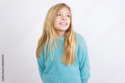Cute Caucasian kid girl wearing blue knitted sweater against white wall with thoughtful expression, looks away keeps hands down bitting his lip thinks about something pleasant.