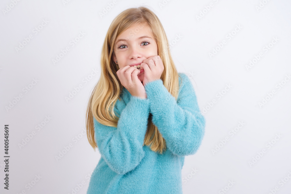 Anxiety - Cute Caucasian kid girl wearing blue knitted sweater against white wall covering his mouth with hands scared from something or someone bitting nails. Anxiety - Cute Caucasian kid girl wearing blue knitted sweater against white wall covering his mouth with hands scared from something or someone bitting nails.