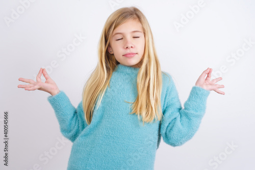 Cute Caucasian kid girl wearing blue knitted sweater against white wall doing yoga, keeping eyes closed, holding fingers in mudra gesture. Meditation, religion and spiritual practices. photo