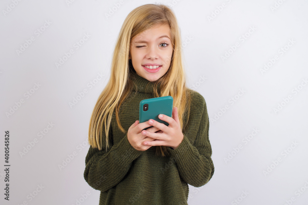 Caucasian kid girl wearing green knitted sweater against white wall taking a selfie celebrating success Caucasian kid girl wearing green knitted sweater against white wall taking a selfie celebrating success