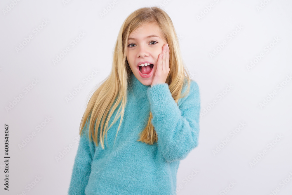 Shocked, astonished Cute Caucasian kid girl wearing blue knitted sweater looking surprised in full disbelief wide open mouth with hand near face. Positive emotion facial expression body language. Shocked, astonished Cute Caucasian kid girl wearing blue knitted sweater looking surprised in full disbelief wide open mouth with hand near face. Positive emotion facial expression body language.