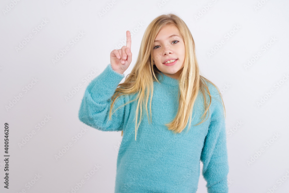Cute Caucasian kid girl wearing blue knitted sweater against white wall showing and pointing up with fingers number one while smiling confident and happy. Cute Caucasian kid girl wearing blue knitted sweater against white wall showing and pointing up with fingers number one while smiling confident and happy.