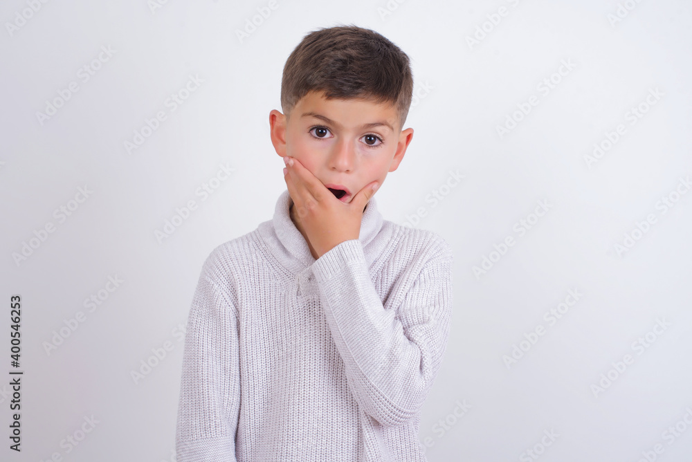 Emotional Cute Caucasian kid boy wearing knitted sweater against white wall gasps from astonishment, covers opened mouth with palm, looks shocked at camera. Reaction concept Emotional Cute Caucasian kid boy wearing knitted sweater against white wall gasps from astonishment, covers opened mouth with palm, looks shocked at camera. Reaction concept