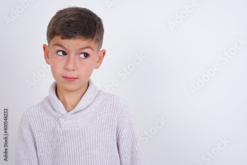 Amazed Cute Caucasian kid boy wearing knitted sweater against white wall bitting lip and looking tricky to empty space.