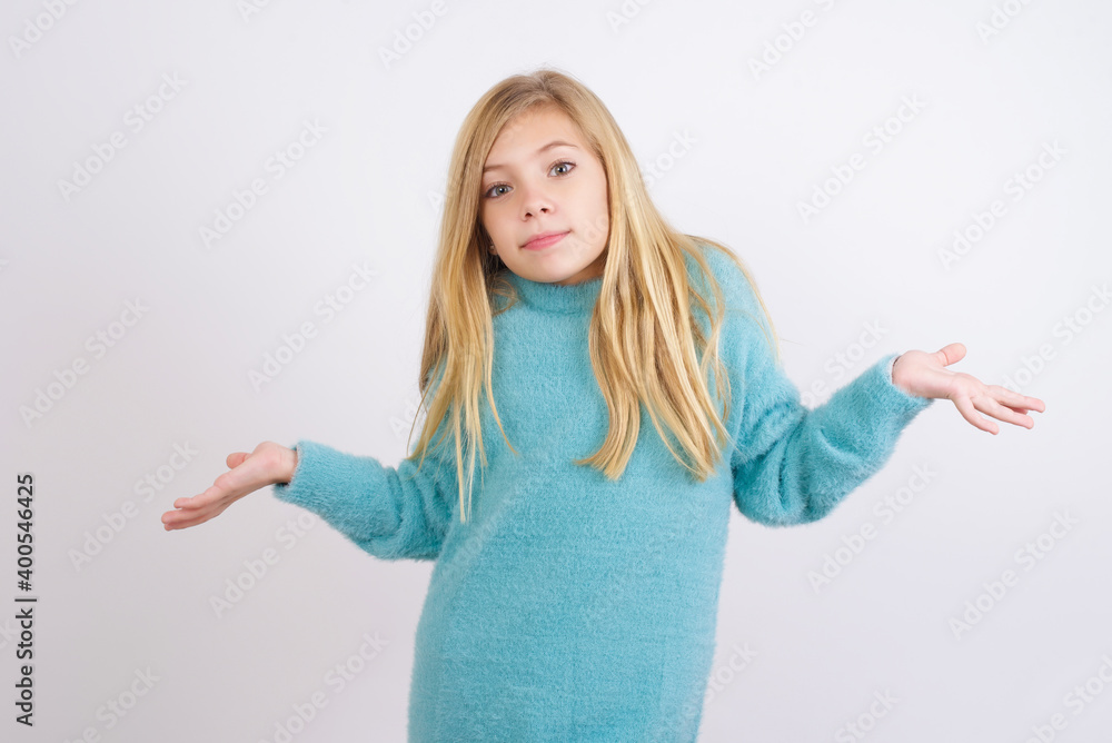 Puzzled and clueless Cute Caucasian kid girl wearing blue knitted sweater against white wall with arms out, shrugging shoulders, saying: who cares, so what, I don't know. Negative human emotions. Puzzled and clueless Cute Caucasian kid girl wearing blue knitted sweater against white wall with arms out, shrugging shoulders, saying: who cares, so what, I don't know. Negative human emotions.