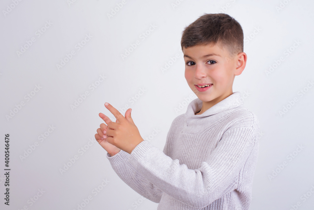 Cute Caucasian kid boy wearing knitted sweater against white wall indicating finger empty space showing best low prices, looking at the camera Cute Caucasian kid boy wearing knitted sweater against white wall indicating finger empty space showing best low prices, looking at the camera
