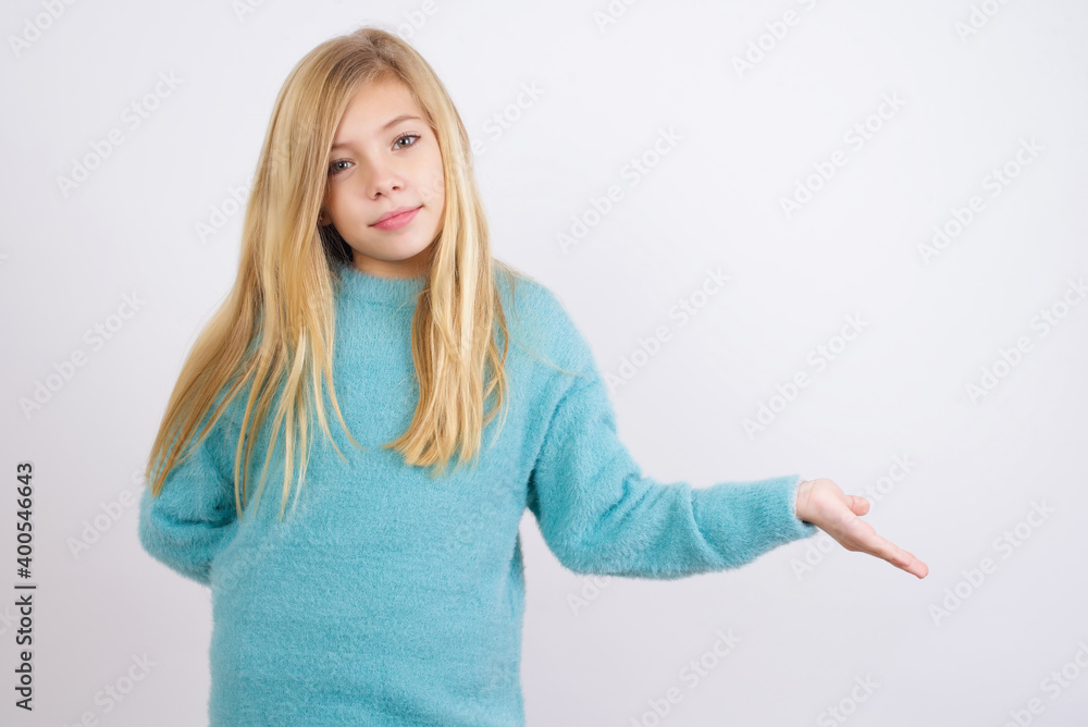 Portrait of Cute Caucasian kid girl wearing blue knitted sweater against white wall with arm out in a welcoming gesture. Portrait of Cute Caucasian kid girl wearing blue knitted sweater against white wall with arm out in a welcoming gesture.
