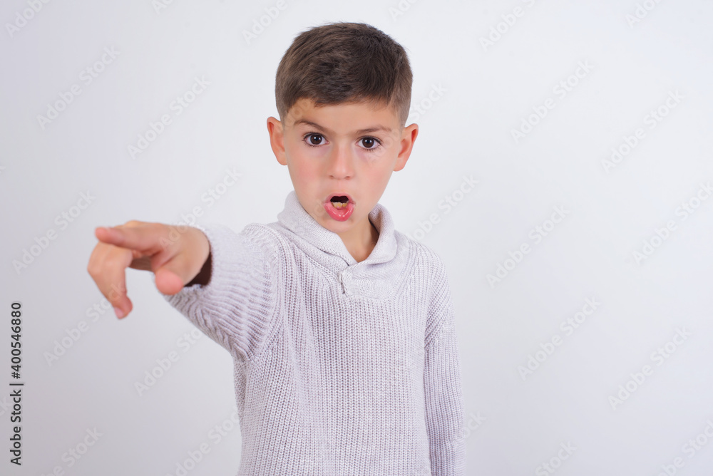 Cute Caucasian kid boy wearing knitted sweater against white wall Pointing with finger surprised ahead, open mouth amazed expression, something on the front. Cute Caucasian kid boy wearing knitted sweater against white wall Pointing with finger surprised ahead, open mouth amazed expression, something on the front.