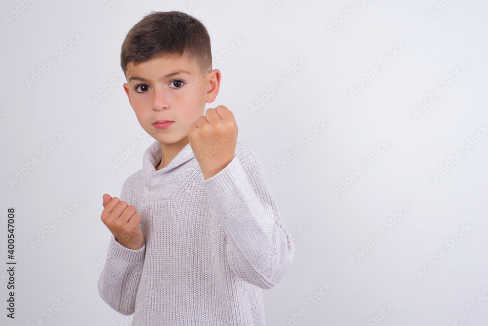 Cute Caucasian kid boy wearing knitted sweater against white wall Ready to fight with fist defense gesture, angry and upset face, afraid of problem. Cute Caucasian kid boy wearing knitted sweater against white wall Ready to fight with fist defense gesture, angry and upset face, afraid of problem.